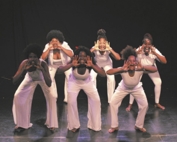 Fotografia de um grupo de mulheres em um palco, uniformizadas com roupas brancas, enfileiradas em duas linhas de três mulheres cada. Todas elas estão com os joelhos flexionados e o tronco inclinado para a frente, com as mãos abertas ao lado da boca.