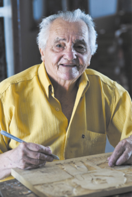 Fotografia de J. Borges. Ele é um homem idoso, com cabelos grisalhos, vestido com uma camisa. Ele está sentado com uma goiva na mão, entalhando uma placa de madeira sobre uma mesa.