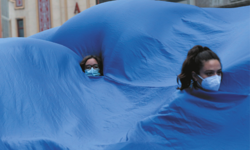 Fotografia de pessoas com as cabeças para fora de um tecido azul que cobre todo o corpo delas. Elas estão usando máscaras de proteção facial, em um protesto em uma via pública.