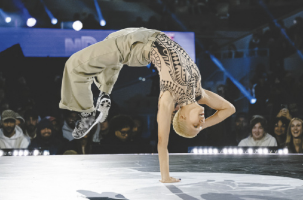 Fotografia de Maia. Ela é uma mulher com cabelos curtos descoloridos, executando um movimento acrobático de breaking em um palco, com a cabeça para baixo, apoiada somente em uma das mãos, com o corpo no ar e as pernas flexionadas.