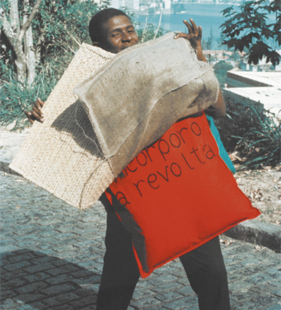 Fotografia de Nildo da Mangueira em pé em uma rua com piso de pedra, com a paisagem do Rio de Janeiro ao fundo. Ele é um homem com cabelos curtos, usando um parangolé feito de camadas de tecidos diversos, que cobrem o tronco e as costas dele. Ele levanta duas camadas do parangolé, mostrando uma almofada em que está escrita a mensagem 'incorporo a revolta'.