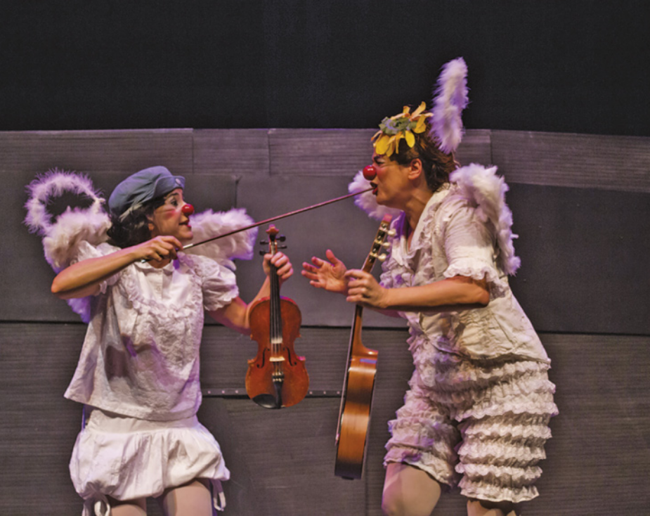 Fotografia de Vera Abbud e Paola Musatti em um palco vestidas como palhaças, fantasiadas como anjos, com asas plumadas nas costas. Vera Abbud está com um violino na mão e enfia o arco do violino na boca de Paola Musatti, que está segurando um pequeno violão.