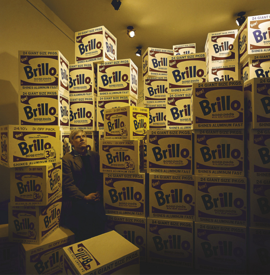 Fotografia de Andy Warhol: um homem com cabelos louros em pé em meio a pilhas de caixas de formato quadrado com o rótulo da embalagem da marca Brillo impresso nas faces das caixas.