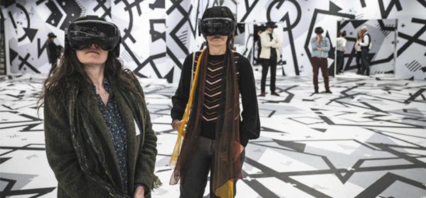 Fotografia de duas mulheres usando óculos de realidade virtual em uma sala expositiva em que as paredes e o piso estão cobertos de estampas gráficas de formas geométricas cinzas e pretas sobre um fundo branco.