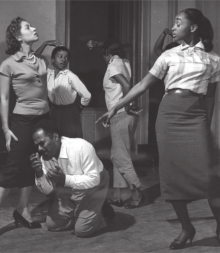 Fotografia em preto e branco de um grupo de atrizes em pé em um cenário teatral, uma delas apontando o dedo indicador para um ator que está ajoelhado no chão, com expressão de tristeza, levando as mãos em direção ao rosto.