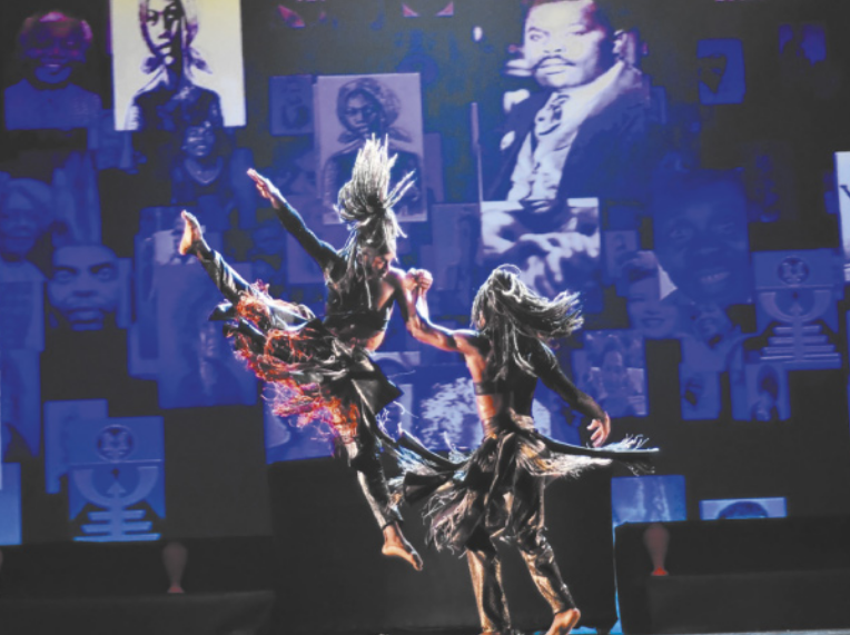 Fotografia de dois bailarinos em um palco, vestidos com saias e trajes inspirados em culturas tradicionais africanas. Um deles está saltando no ar, com as pernas afastadas e estendidas, e o outro bailarino segura uma das mãos dele. O pano de fundo do cenário mostra diversos retratos de personalidades negras, além de objetos simbólicos de culturas afrodescendentes.