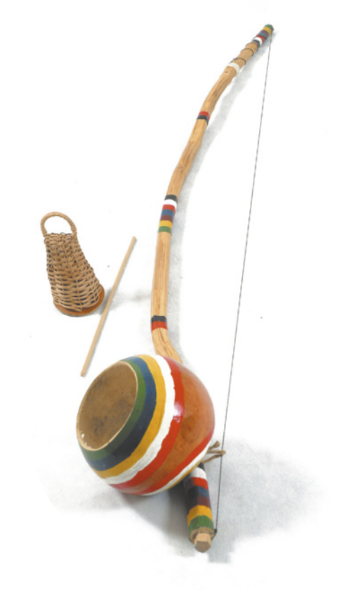 Fotografia de um berimbau: um instrumento com um arco de madeira decorado com faixas coloridas pintadas ao longo dele, e com um fio de arame esticado entre as extremidades do arco. Na parte inferior dele está acoplada uma cabaça com uma abertura. Acompanham o berimbau uma vareta fina de madeira e um chocalho de palha chamado caxixi.