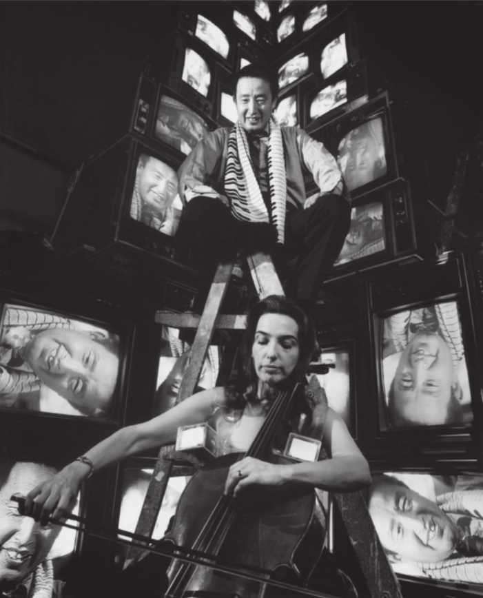 Fotografia em preto e branco de Nam June Paik: um homem com cabelos curtos, usando um cachecol, sentado sobre uma escada de madeira em frente a uma torre feita com televisores ligados mostrando a imagem do rosto de Nam June Paik. A fotografia também mostra Charlotte Moorman. Ela é uma mulher de cabelos longos tocando um violoncelo em frente à instalação e à escada em que Nam June Paik está sentado.