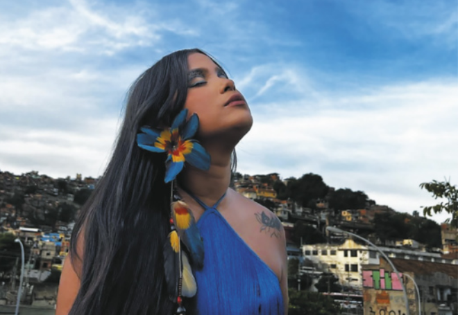 Fotografia de Kaê Guajajara. Ela é uma mulher indígena com cabelos longos e lisos, usando brincos enfeitados com penas de aves. Ela está de olhos fechados, com a cabeça voltada para cima, com uma comunidade de casas em um morro ao fundo.