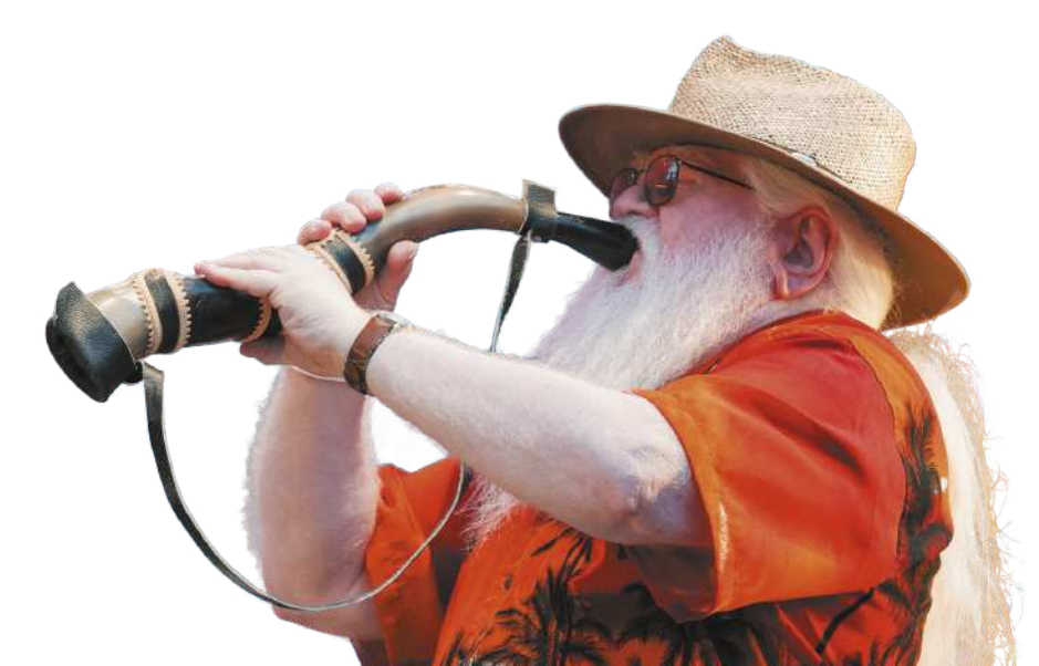 Fotografia de Hermeto Pascoal. Ele é um homem idoso, com a barba e os cabelos grisalhos e longos, de óculos, usando um chapéu e uma camisa de estampa florida. Ele está tocando um berrante, feito com um chifre de boi longo e de formato curvo.