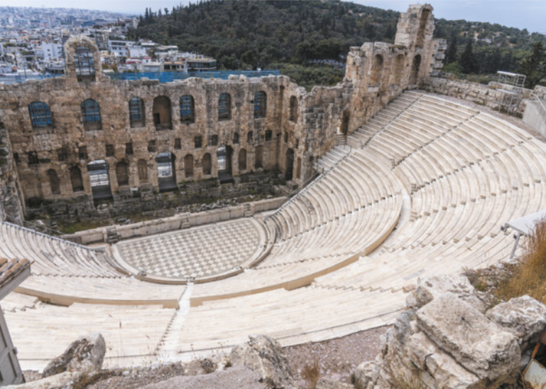 Fotografia de ruínas de um teatro com arquibancadas de formato semicircular, diante de uma arena circular ao centro e de um palco retangular. Ao fundo do palco há um edifício com janelas em forma de arcos. Ao fundo há uma cidade e um morro coberto por vegetação.