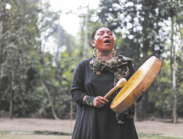 Fotografia de uma mulher indígena cantando e tocando um tambor largo, semelhante a um pandeiro grande, usando uma baqueta. Ela está de vestido preto e usa brincos e um lenço no pescoço. Ao fundo, há uma mata.