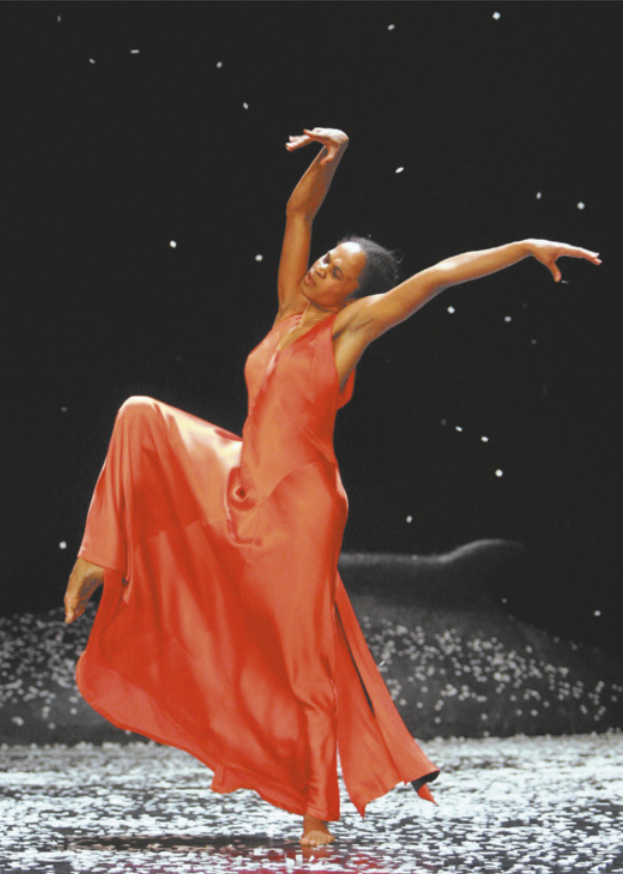 Fotografia de uma bailarina dançando em um palco enquanto flocos de neve artificial caem ao seu redor. Ela usa um vestido longo, vermelho e esvoaçante. Está em pé, com um dos joelhos flexionado e elevado, os braços estendidos, um para o alto e o outro em diagonal, e a cabeça inclinada na mesma direção do braço inferior.