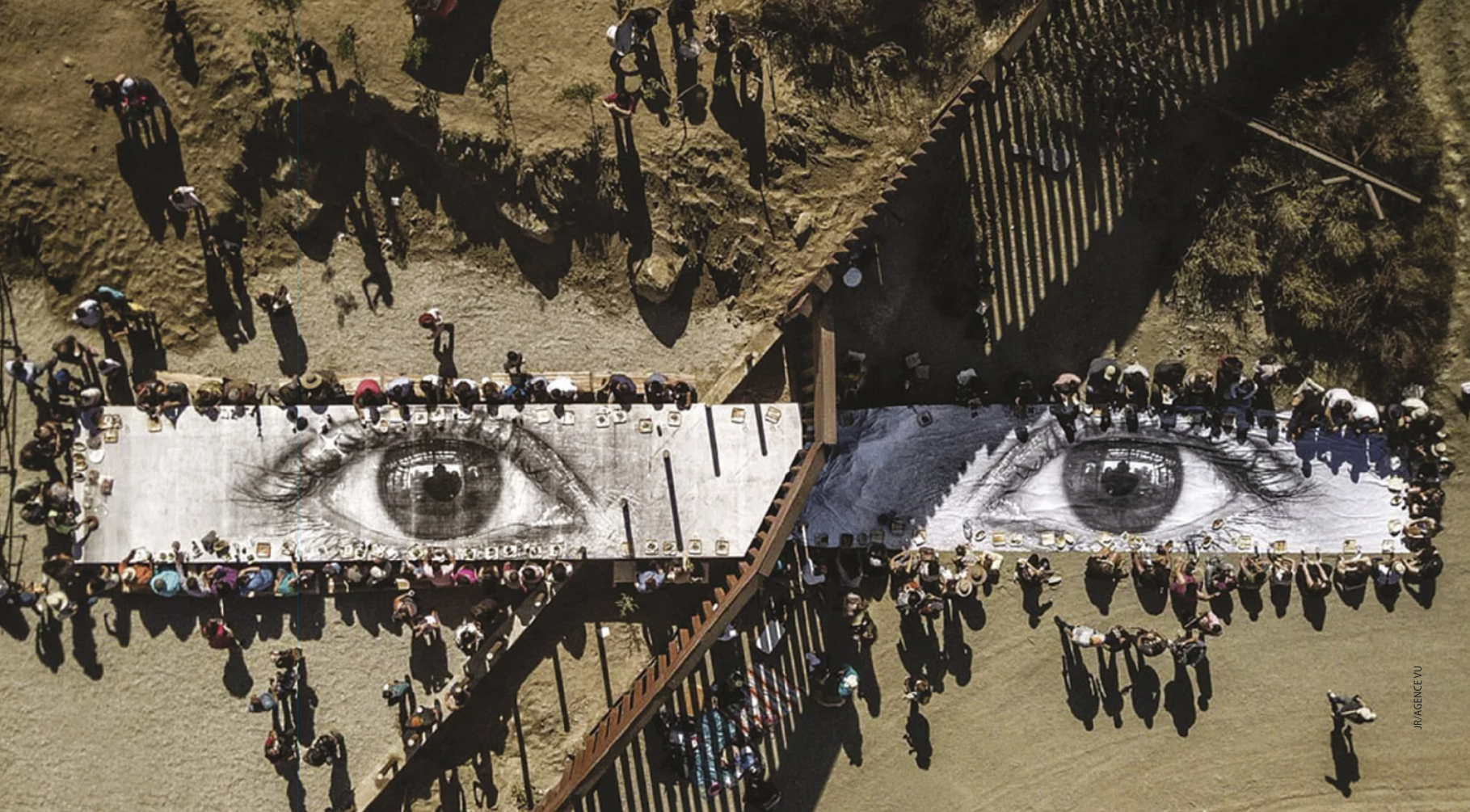 Fotografia aérea com instalação de uma grande faixa de tecido no chão de uma paisagem árida, com a imagem de dois olhos. Há um muro de metal que corta a paisagem, e cada olho da imagem está de um dos lados do muro. De ambos os lados do muro há pessoas sentadas diante da faixa de tecido, com pratos de comida dispostos sobre ela.