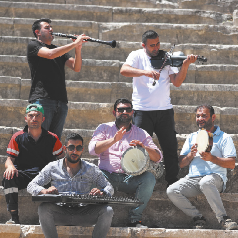 Fotografia de um grupo de homens tocando instrumentos em uma escadaria. Dois deles estão em pé tocando violino e clarinete; e os sentados tocam uma espécie de pandeiro, denominado riqq; um tambor de formato alongado, denominado darbuka; e um instrumento de cordas semelhante a uma cítara, que é tocado apoiado sobre as pernas do músico, deitado na horizontal, denominado de saltério ou kanun. Um deles está sentado sem instrumentos.