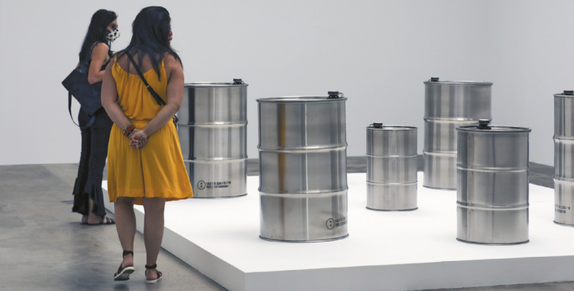 Fotografia de mulheres com máscaras de proteção facial em um espaço expositivo, diante de uma instalação com tambores de metal cilíndricos, de tamanhos diversos, dispostos sobre um pedestal no chão. Na tampa de cada tonel há um pequeno aparelho eletrônico instalado.