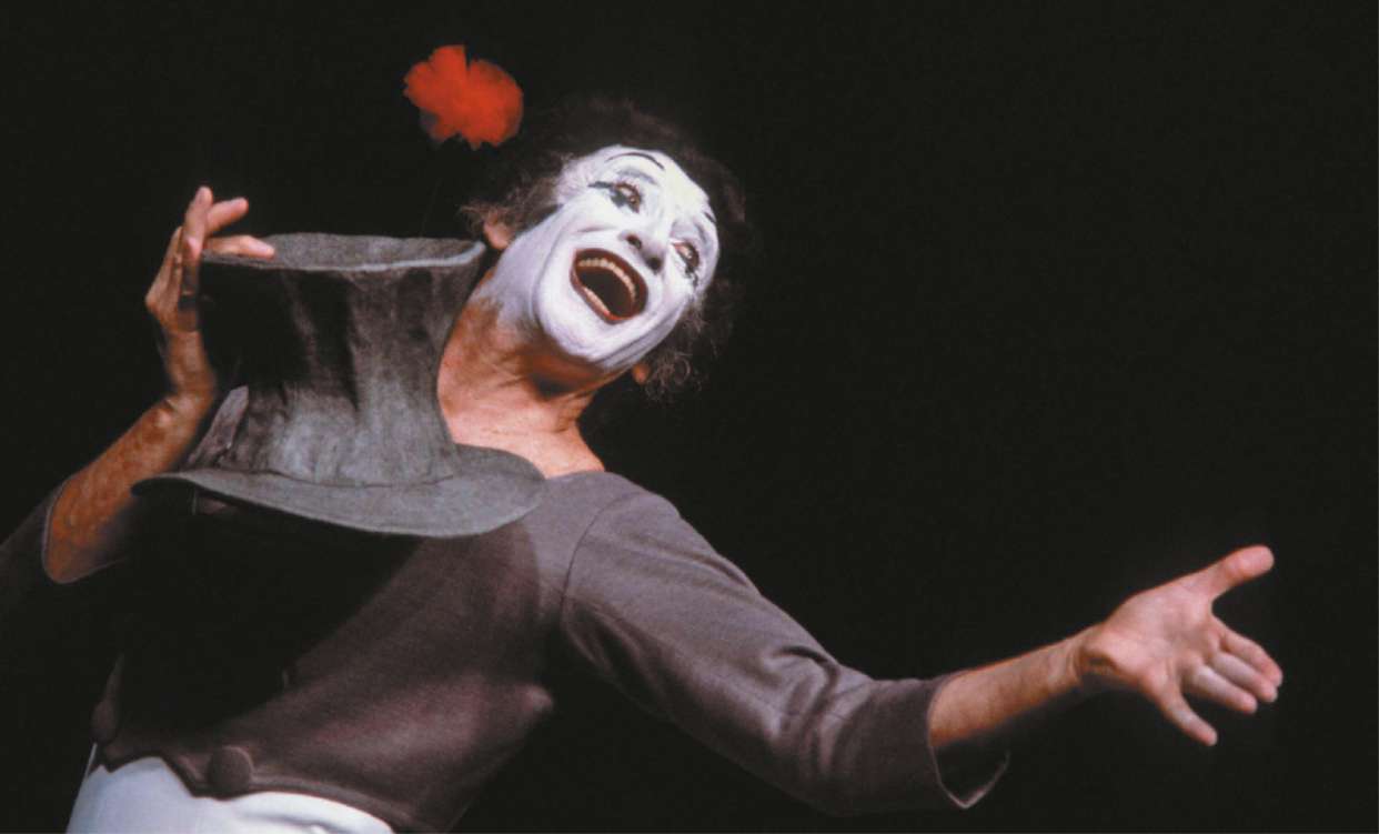 Fotografia de Marcel Marceau. Ele é um homem com cabelos grisalhos e cacheados, vestido como palhaço, segurando uma cartola e sorrindo com o corpo inclinado para trás, estendendo a mão para o lado.