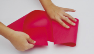 Fotografia de mãos dobrando uma metade de folha de papel colorida, de formato alongado.