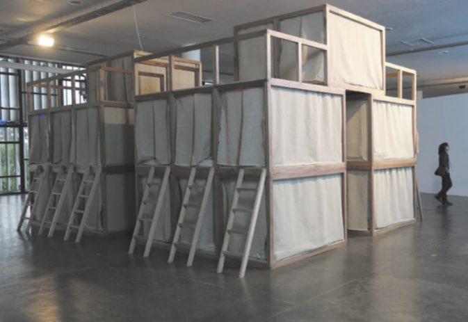 Fotografia de instalação em um espaço expositivo com instalação artística de estruturas de madeira revestidas com tecidos, configurando pequenos espaços compartimentados por divisórias de tecido, denominados de 'ninhos'. Há escadas de madeira que dão acesso aos ninhos, e ao fundo há uma mulher caminhando ao lado da instalação.