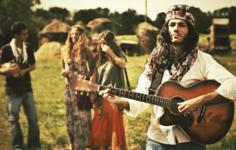 Fotografia de jovens com trajes da moda hippie, com cabelos longos, faixas nos cabelos e roupas leves com estampas floridas. Um jovem está tocando um violão, e atrás dele há duas garotas e um garoto conversando, em uma paisagem rural.