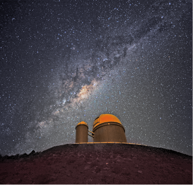 Fotografia noturna com destaque para o céu com muitos pontos luminosos, as estrelas e ao centro uma região mais iluminada, a via Láctea. Abaixo duas construções com o teto arredondado e o solo rochoso.