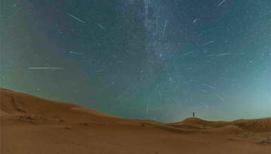 Fotografia de uma paisagem, com o céu escuro com traços e pontos luminosos. Abaixo está o solo arenoso.