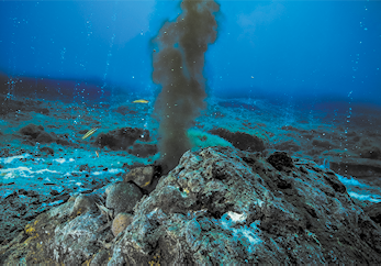 Fotografia de um aglomerado de rochas no fundo do mar, com água saindo de uma fissura, com coloração escura e aspecto de fumaça.