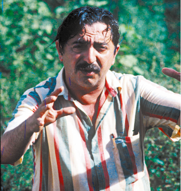Fotografia de um homem de cabelos curtos e pretos, com franja na testa e aspecto de molhado. Ele tem bigode, e está com a testa franzida. Seus braços estão levantados para frente e as mãos abertas. Ele usa uma camisa com listras coloridas e ao fundo, há vegetação.