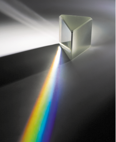Fotografia de um ambiente escuro, com um prisma triangular no centro. À esquerda, um feixe de luz branca atinge o prisma. À frente, saem feixes de luz colorida.