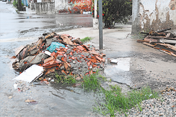 Fotografia de uma esquina cujas ruas estão alagadas. Há entulho com restos de construção na rua e em cima da calçada. Ao lado há ripas de madeira jogadas na calçada e encostadas em parte de uma parede.