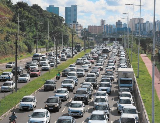 Fotografia de duas avenidas separadas por um canteiro de grama e congestionada por inúmeros carros, além de motos, caminhões e ônibus. No canteiro central e na lateral direita de uma das avenidas há postes de luz e à esquerda há um paredão formado por um morro com muita vegetação. Ao fundo há diversos prédios de uma cidade e parte do céu que está claro.