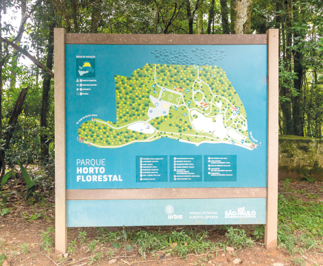 Fotografia de uma placa presa, no chão de terra, por duas estacas de madeira. Ao fundo árvores de uma floresta. Na placa há os dizeres: Parque Horto Florestal e há uma cartografia com pontos amarelos e verdes que representam vegetação, espaços em branco representando caminhos e construções e símbolos de localização. Abaixo da cartografia, há uma legenda.