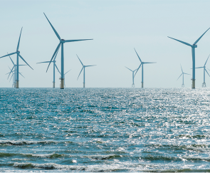 Fotografia de parte do oceano com usina eólica offshore composta de várias torres espaçadas, com um gerador e 3 pás em cada uma.