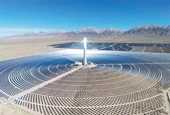 Fotografia aérea de uma grande área repleta de placas solares dispostas uma ao lado da outra formando vários círculos com espaçamento entre eles e no centro uma torre receptora da radiação solar. Ao fundo há uma cadeia de montanhas rochosas.
