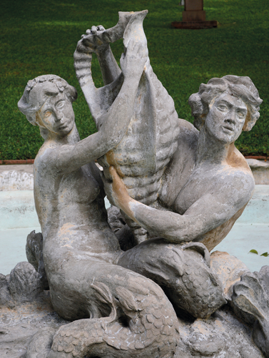 Fotografia de um chafariz composto pela estátua de uma mulher e de um homem, ambos com caudas de peixe ao invés de pernas e segurando juntos uma concha marinha.