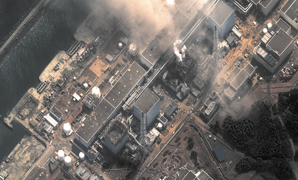 Fotografia aérea mostrando uma usina nuclear por cima com vários prédios e instalações e muita fumaça escura saindo de um dos prédios.