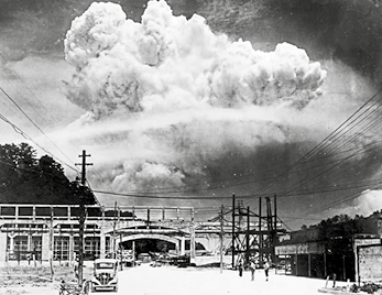 Fotografia em preto e branco de uma rua com construções ao fundo e uma grande nuvem de fumaça ocupando o céu.