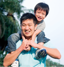 Fotografia de um homem oriental, visto da cintura para cima. Ele tem cabelos pretos e usa camiseta de manga curta. Em seus ombros, há um menino com cabelos pretos, também oriental, de camiseta de manga curta, calça comprida, tênis e, sentado passando seus braços ao redor da cabeça do homem e ambos estão dando as mãos e sorrindo.