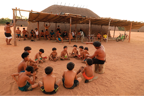 Fotografia de várias crianças indígenas sentadas em roda no chão e junto delas há um homem, também indígena sentado em um tronco gesticulando com a mão. Eles estão em um chão de areia e ao fundo há vários adultos.