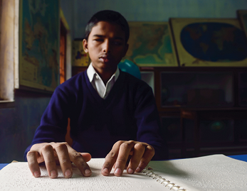 Fotografia de um menino sentado em frente a uma mesa que possui um livro em sistema braile. Ele está com as duas mãos passando sobre o livro.