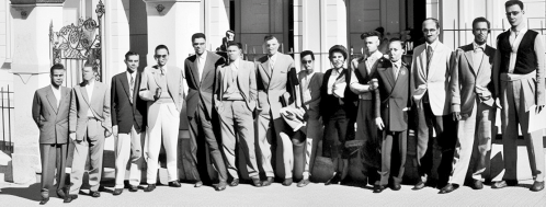 Fotografia em preto e branco de 13 homens e 1 mulher. Todos estão em pé um ao lado do outro à frente de um edifício e a maioria está usando terno.