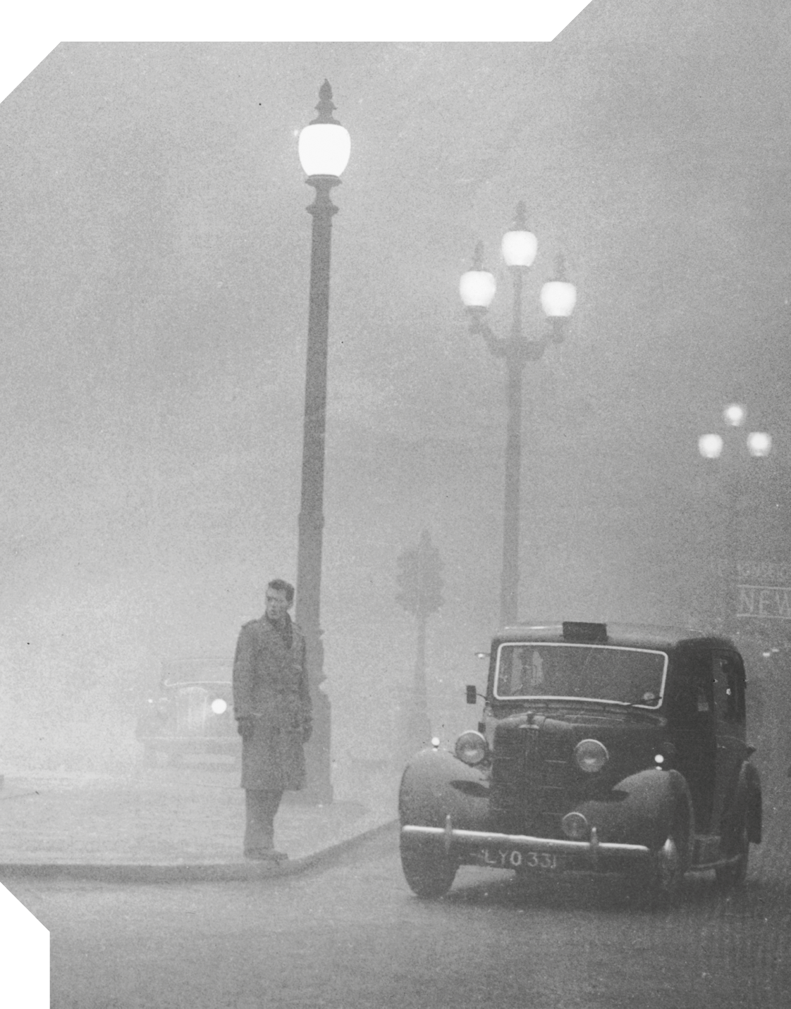 Fotografia em preto e branco de uma rua com um carro antigo e, à esquerda, um homem com calça e casaco comprido no limite da calçada e próximo a um poste com a luz acesa. Há muita névoa esbranquiçada, outro carro e dois postes ao fundo.