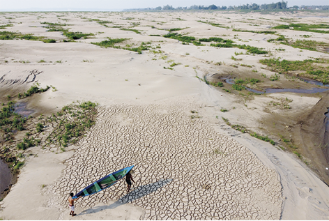 Fotografia da vista aérea de uma região arenosa com rachaduras entremeadas por uma vegetação rasteira. Na margem inferior estão duas pessoas carregando um barco.