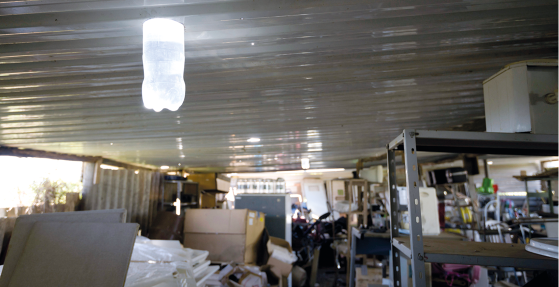 Fotografia de um ambiente interno iluminado, com vários móveis e objetos espalhados. No teto, há uma garra pet transparente com um líquido transparente acoplada ao telhado. A garrafa está iluminada.