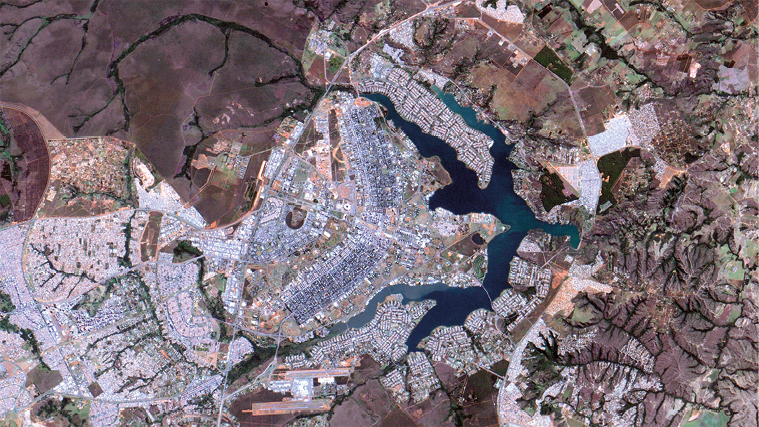 Fotografia aérea de satélite de uma cidade. Ao centro há um corpo d’água maior e no entorno, outros corpos d’água menores e vegetação. No entorno do corpo d’água maior, há áreas de construções em formato retangular.