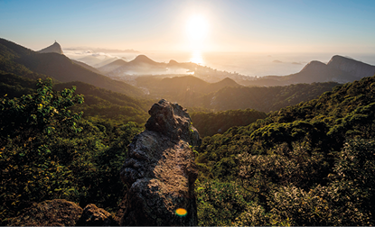 Fotografia de uma área montanhosa com vegetações, e ao fundo montanhas e sobre elas, o céu claro com Sol forte nascendo.