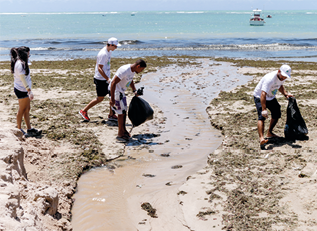 Fotografia de quatro pessoas caminhando pela areia em uma praia. Elas estão recolhendo resíduos do chão e segurando sacos de lixos.