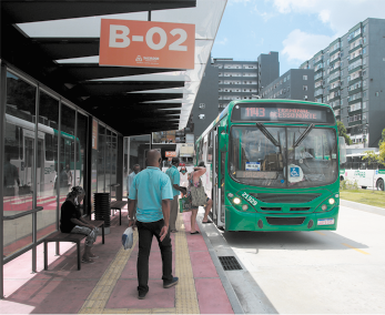 Fotografia de pessoas em um ponto de terminal de ônibus. O local tem uma cobertura de proteção e bancos, e na rua, um ônibus. No entorno há construções.
