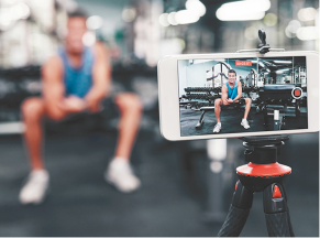 Fotografia, com um celular sobre um tripé em primeiro plano. O celular está filmando uma pessoa se exercitando em uma academia. Em segundo plano, desfocado, está a pessoa se exercitando.
