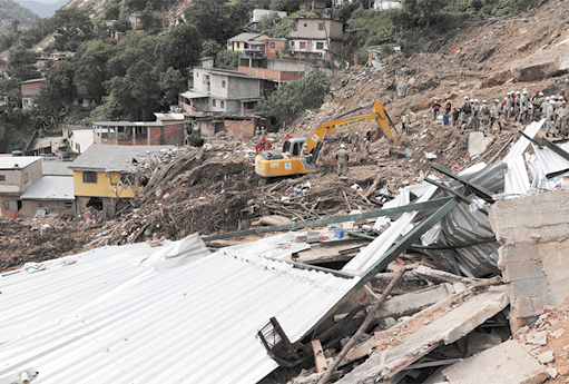 Fotografia de uma área de declive com muitos entulhos partes de construções destruídas. Há uma escavadeira no local e várias pessoas com capacetes. Ao fundo há construções residenciais.